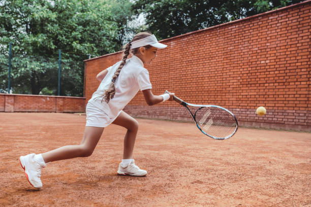 Child playing tennis on outdoor clay court. Full length shot of a tanned little girl on tennis court. Girl child playing tennis against the wall. Summer activities for children.